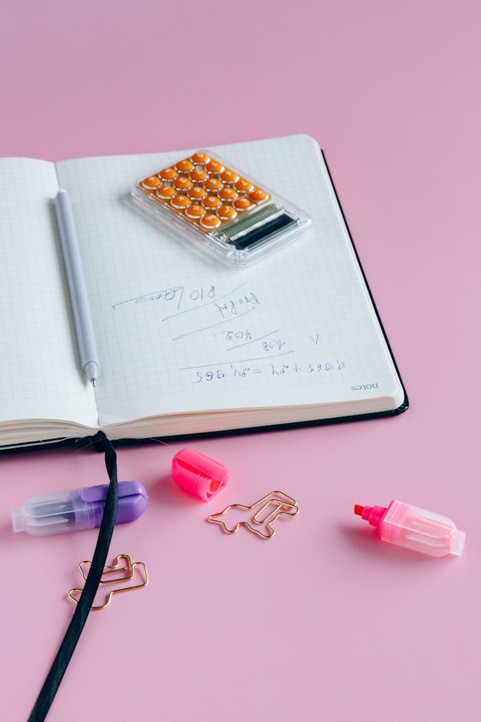 A notebook with calculations, pens, and items on a pink background.