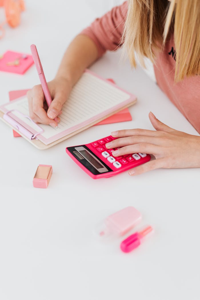 A woman writing in a notepad with a pink calculator nearby, ideal for education and finance themes.